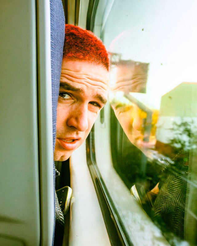 A person with short, bright red hair leans against a train window, looking outside. Their face is partially reflected in the glass, and sunlight streams in, illuminating their expression.
