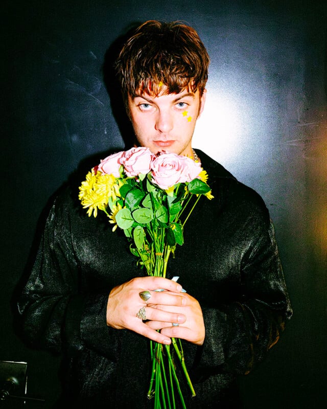 A person with short brown hair and a star sticker on their cheek holds a bouquet of pink and yellow flowers, standing against a dark background while looking directly at the camera.