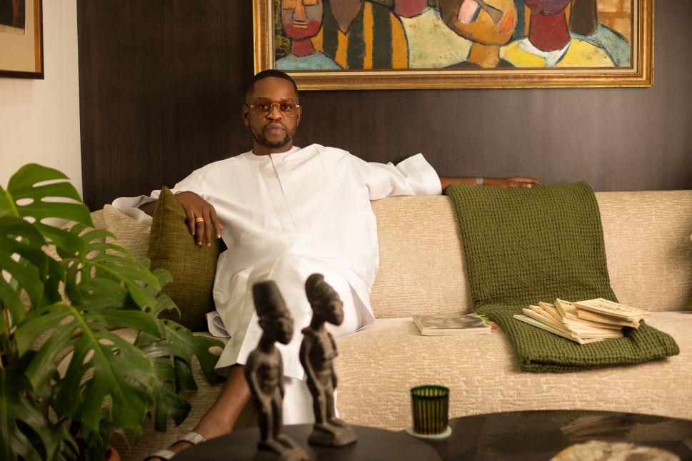 A portrait shows Nigerian lawyer, collector and cultural entrepreneur Kayode Adegbola seated on a cream sofa in a living room, dressed in a white outfit with green accents around him and artworks visible on the wall behind.