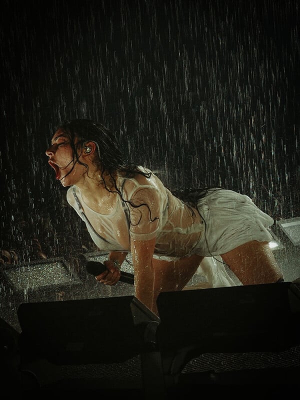 A performer in a wet white outfit crouches dramatically on stage, gripping a microphone and singing passionately in heavy rain.