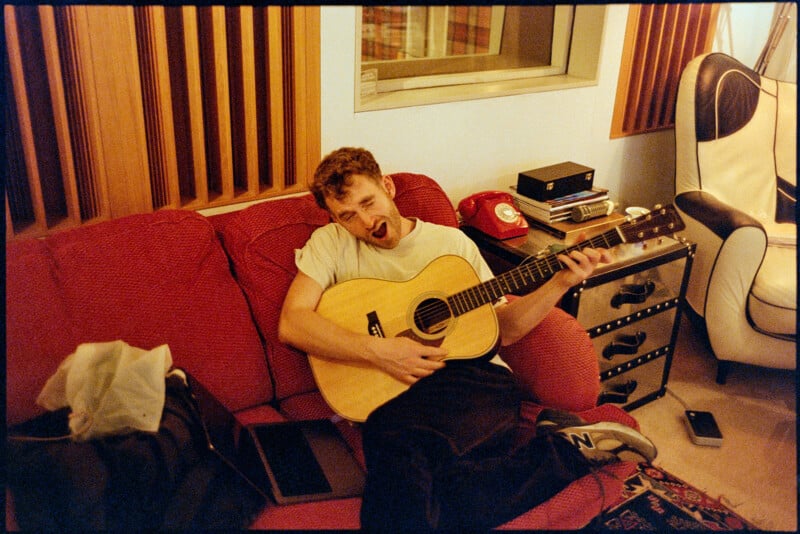 A man sits on a red couch playing an acoustic guitar, making a funny face. Around him are a laptop, a phone, and a tissue box. The room has wooden panels, a white chair, and a red rotary phone on a side table.