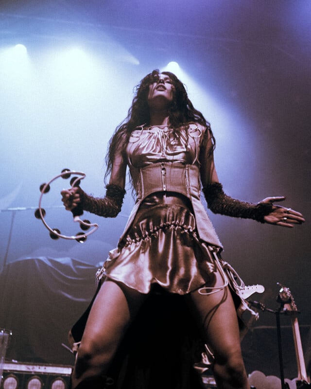 A performer wearing a metallic dress and arm warmers plays a tambourine on stage under dramatic blue and purple lighting.