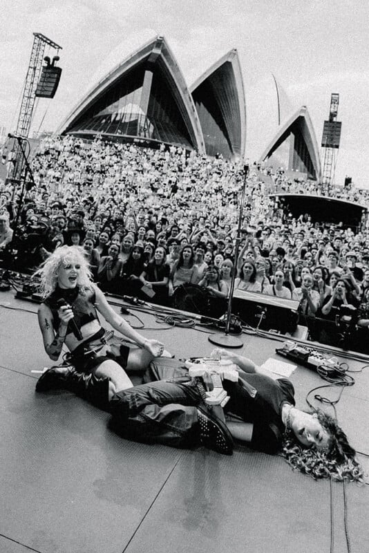 Two musicians perform on an outdoor stage in front of a large crowd, with one kneeling and the other lying down. The Sydney Opera House is visible in the background. The image is in black and white.