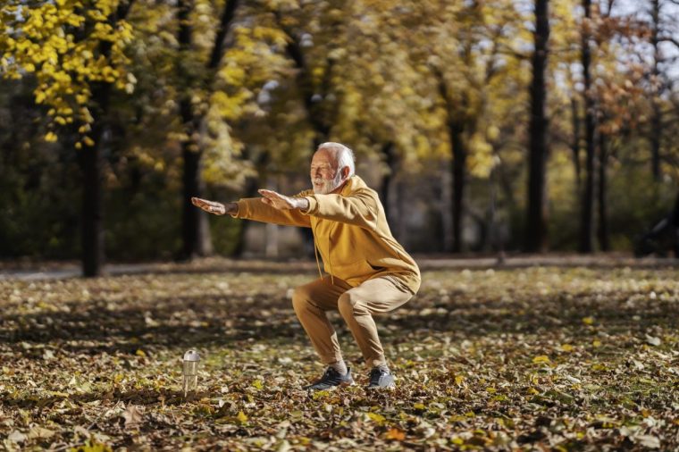 Full length of senior active sporty man in tracksuit doing squats in nature during his fitness day.