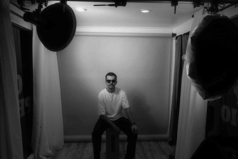 A man wearing sunglasses and a white shirt sits on a stool in the center of a photo studio with professional lighting equipment and a backdrop surrounding him. The image is in black and white.