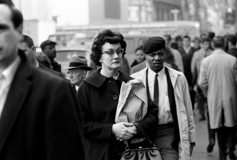A woman in a dark coat and glasses holds a paper bag while walking through a busy city street filled with people, some in hats and coats; the photo is in black and white.