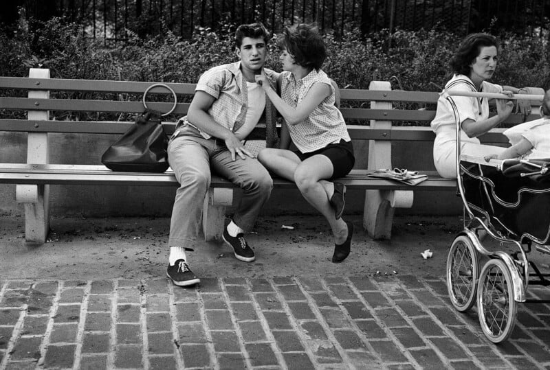 A young couple sits closely on a park bench, the woman touching the man's face as they talk. A large handbag rests beside them. Nearby, a woman in white sits next to a baby stroller, looking away thoughtfully.