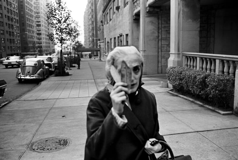 An older woman walks on a city sidewalk, holding a leaf in front of her face, partially covering it. She carries a handbag, and vintage cars and tall buildings line the street in the background.