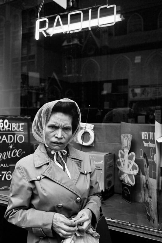 A woman wearing a headscarf and trench coat stands in front of a shop window displaying radios and a neon “RADIO” sign. She looks directly at the camera with a serious expression.