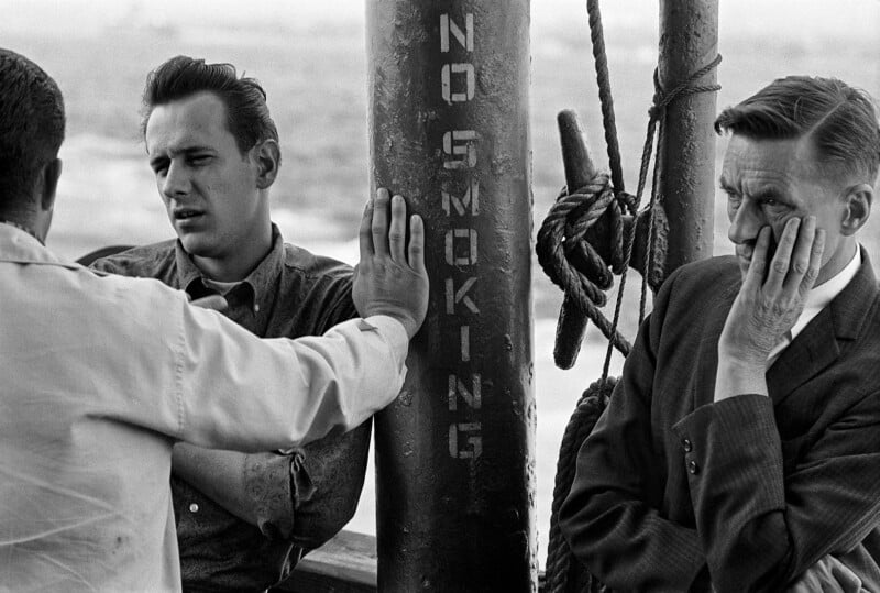 Three men stand near a pole marked "NO SMOKING." One man faces away, gesturing with his hand on the pole; another leans, looking thoughtful; the third man rests his head on his hand, appearing concerned or pensive.