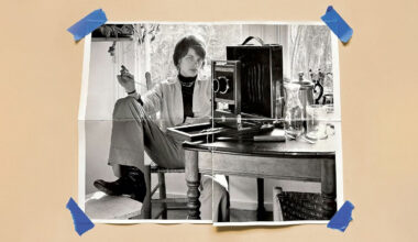 A black-and-white photo of a person sitting casually at a table with a camera and glass pitcher, taped at the corners to a beige surface with blue tape. The person is holding a cigarette and looking at the camera.