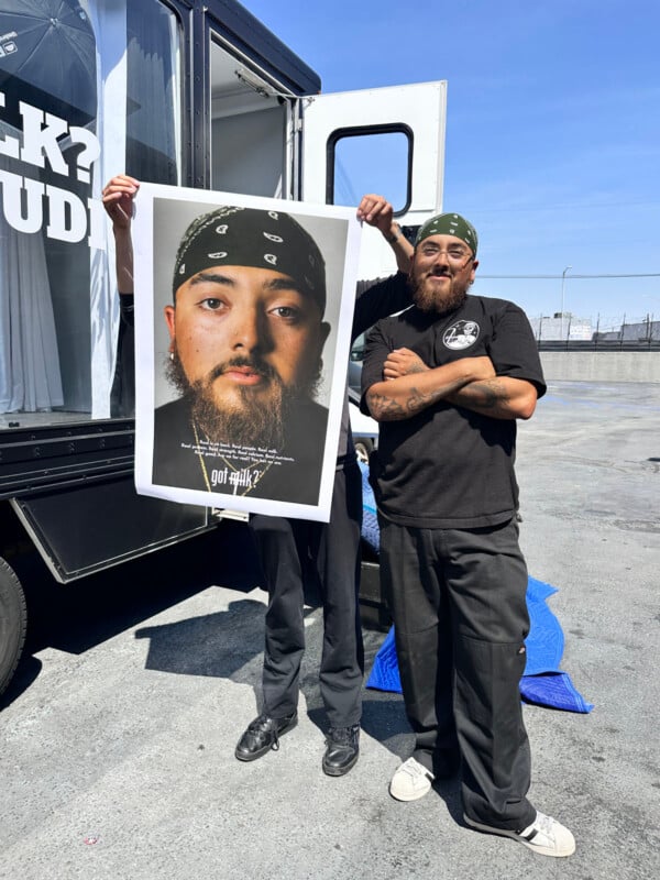 Two men stand outside; one holds a large poster of a man wearing a black bandana and a serious expression. The other man, also in a bandana, black shirt, and pants, stands smiling with arms crossed next to him.