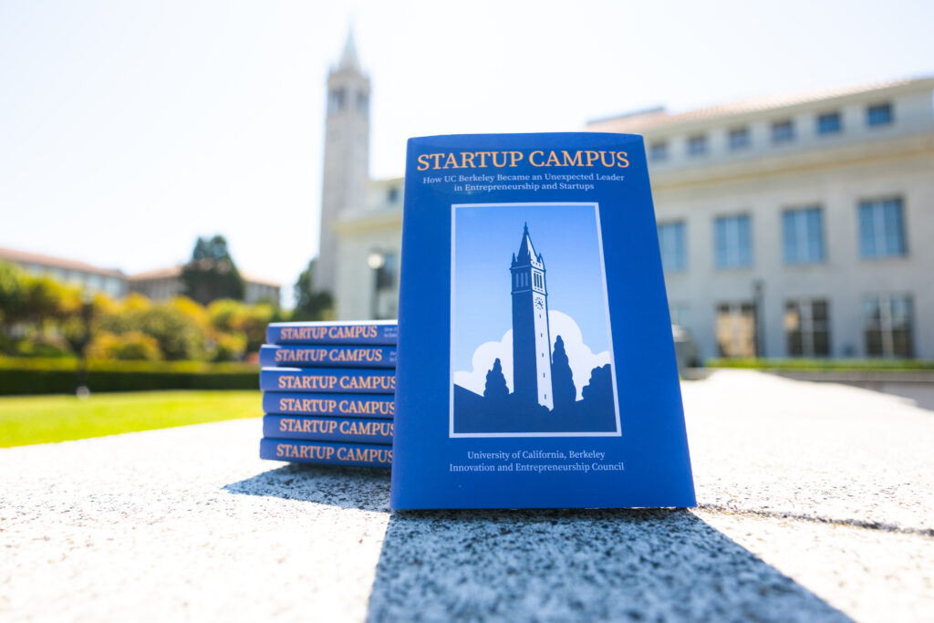 A copy of the Startup Campus book leans against a stack of six copies, with the Campanile blurred in the background
