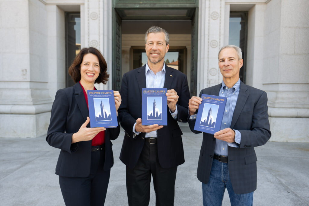 Laura Hassner, Darren Cooke and Mike Alvarez Cohen stand outside a building, each holding in front of them a copy of the Startup Campus book while smiling toward the camera.