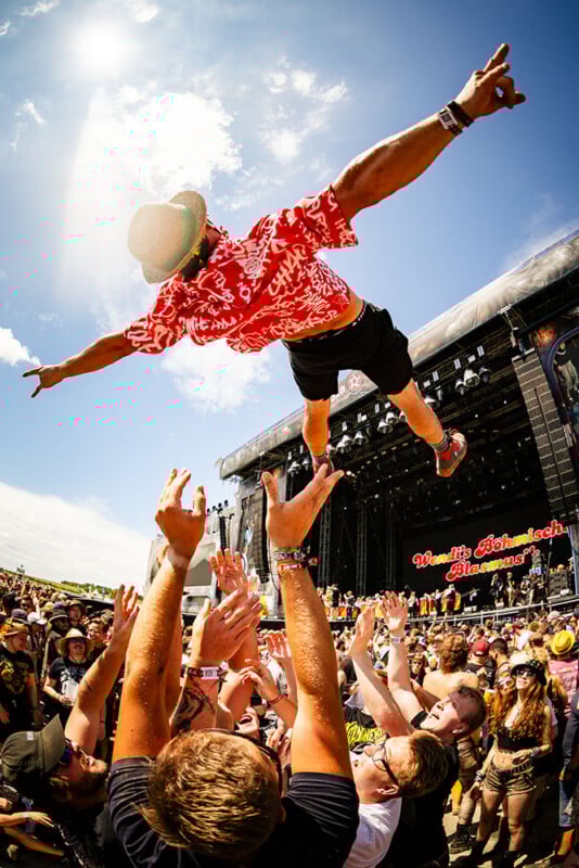 A man in a red shirt and hat is crowd surfing above a cheering audience at an outdoor music festival, with a large stage and bright blue sky in the background.