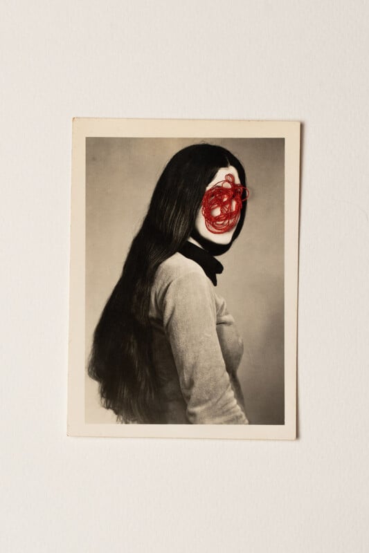 A vintage black-and-white photo of a woman with long dark hair; her face is obscured by red thread tangled and stitched over the facial area, creating an abstract effect.