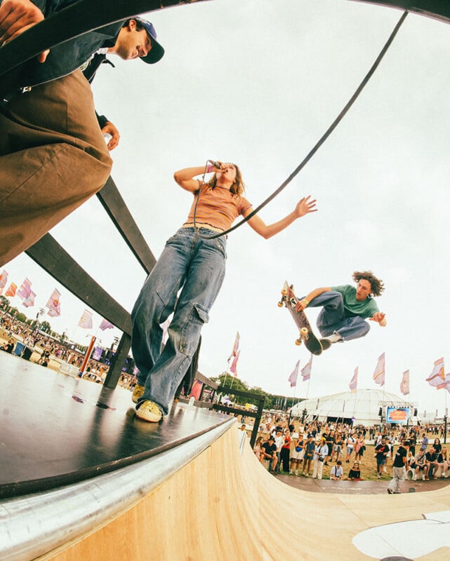A skateboarder performs a high jump trick off a ramp as a woman sings into a microphone and a man sits nearby; a crowd and festival flags fill the background under a cloudy sky.