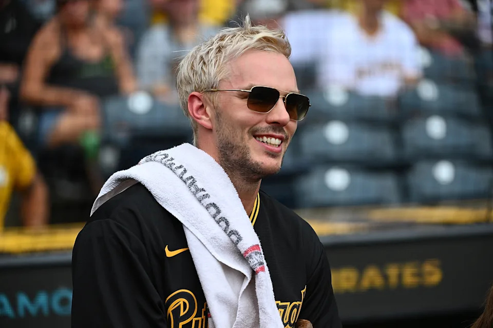 Nicholas Hoult wearing a sports jersey with a towel around the neck, smiling and wearing sunglasses at a sports event