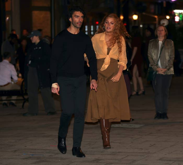Two people walking on a sidewalk at night. The person on the right is wearing a long skirt, knee-high boots, and a loose shirt tied at the waist