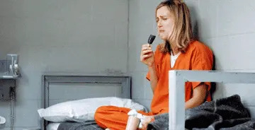Person in an orange prison jumpsuit sits on a bed, appearing upset while eating a snack