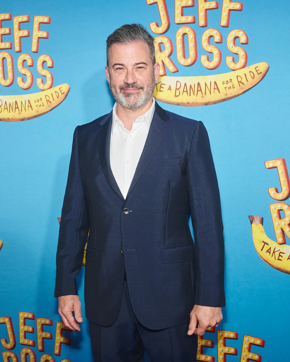A person in a suit poses in front of a backdrop with "Jeff Ross: Take a Banana for the Ride" text and banana illustrations