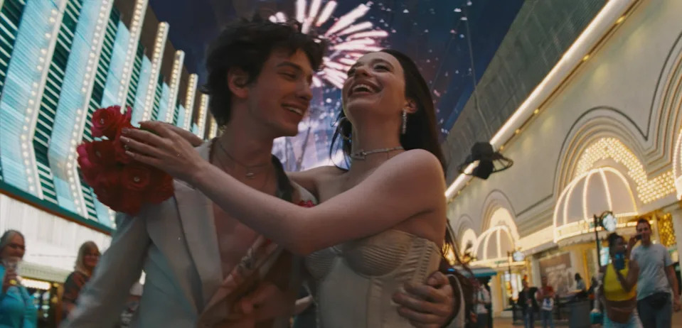 A couple joyfully embrace on a bustling street under fireworks. The woman wears a strapless corset dress; the man is in an open jacket, holding roses