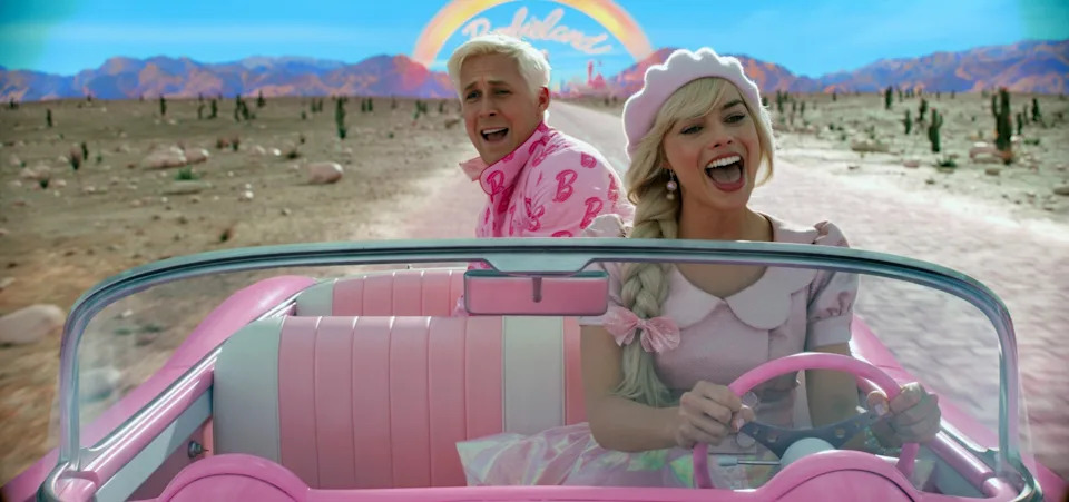 Two people in a pink convertible in a desert, smiling and singing. The passenger wears a patterned shirt, and the driver has a hat and dress
