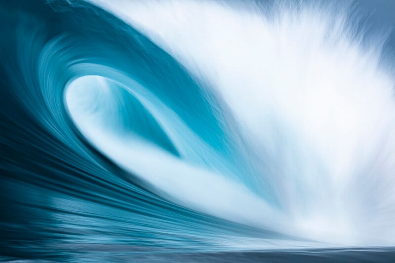 A powerful, bright blue ocean wave curls and crashes, creating a tunnel with white spray and mist against a soft blue sky.