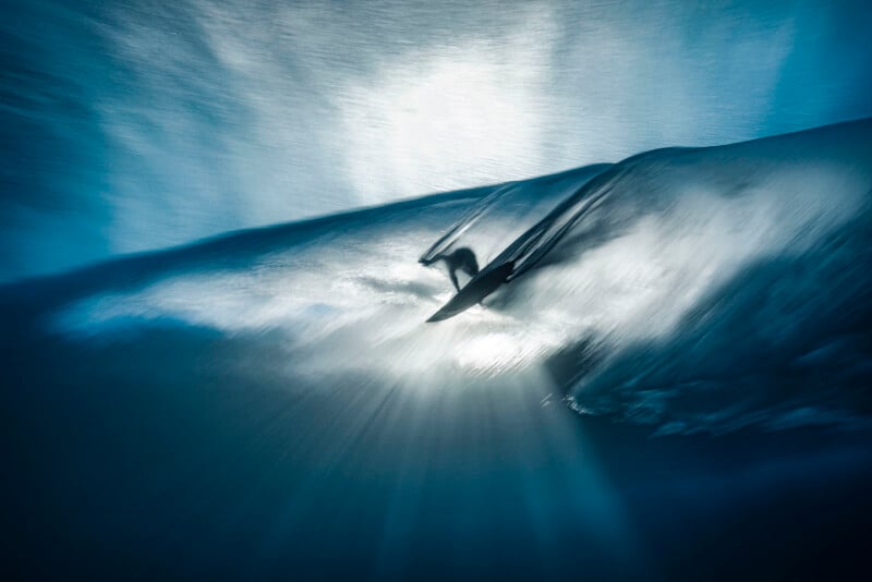A surfer rides inside a powerful wave, illuminated by sunlight streaming through the water, creating a dramatic and dynamic underwater scene with blue tones and motion blur.