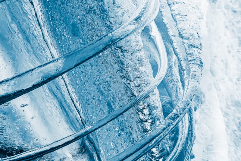 Close-up of curved, clear ice formations with bubbles inside, displaying various shades of blue and white. The texture is smooth and glassy, creating an abstract, chilly appearance.