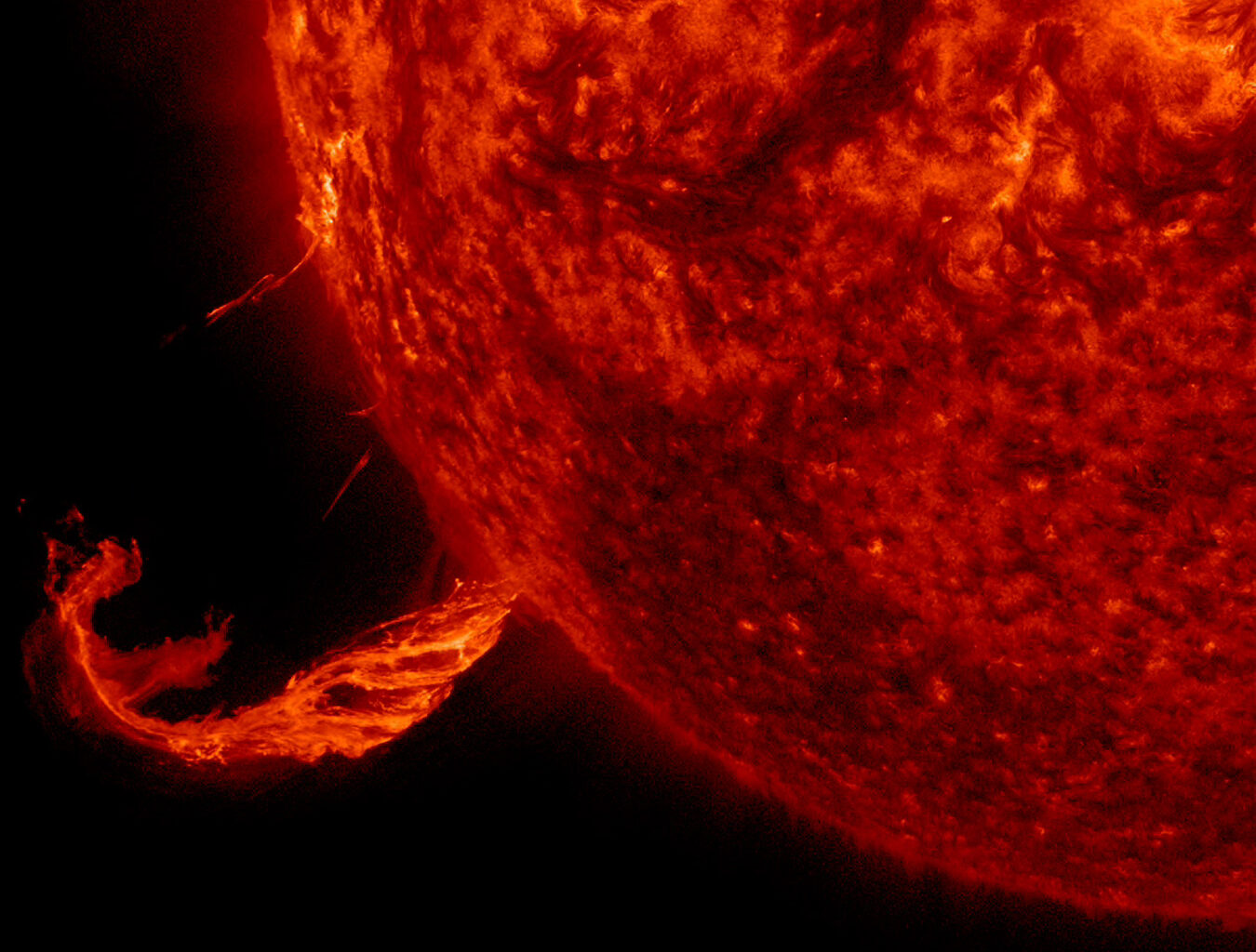 This image focuses on a twisting jet of material erupting from the Sun, glowing bright against the black backdrop of space. The Sun is a fiery, deep red ball taking up three-quarters of the image on the right.