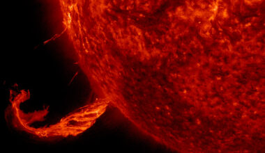 This image focuses on a twisting jet of material erupting from the Sun, glowing bright against the black backdrop of space. The Sun is a fiery, deep red ball taking up three-quarters of the image on the right.