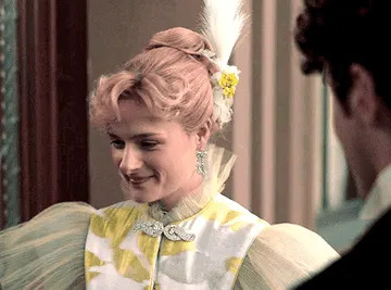 Person in elaborate period costume with puffed sleeves, elegant jewelry, and an intricate hairstyle, smiling at another person