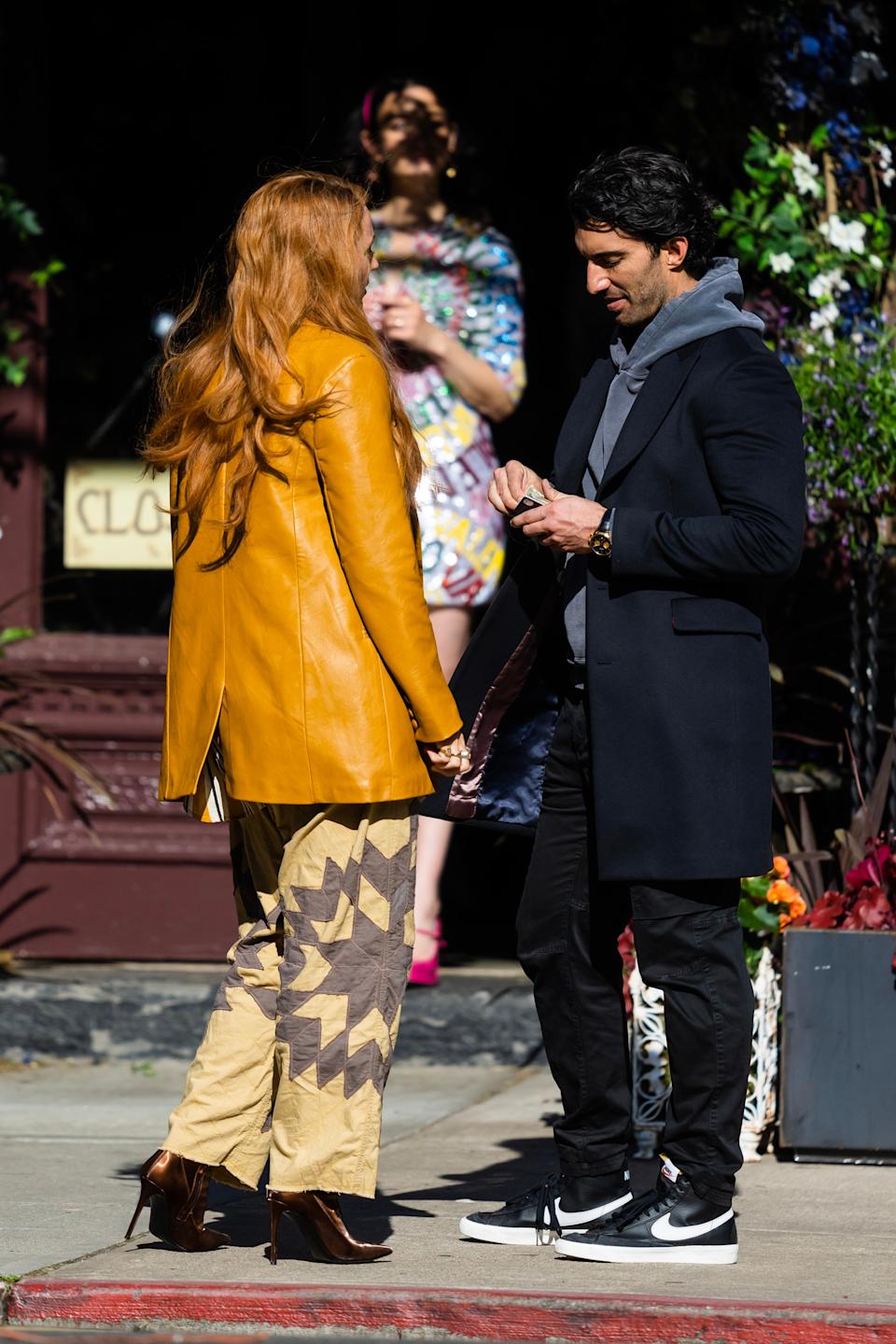 Two people conversing outside; one wears a yellow jacket and patterned pants, the other a dark coat and sneakers. Background figure in colorful attire