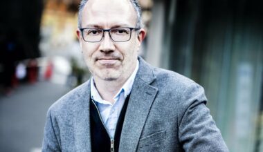 Cale Scharf in a grey blazer, dark blue zip-up sweater, and a white collar shirt beneath, smiling with glasses on
