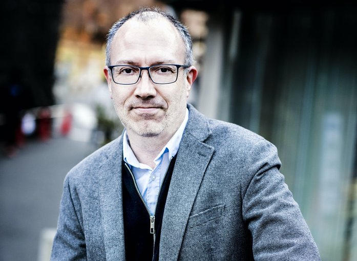 Cale Scharf in a grey blazer, dark blue zip-up sweater, and a white collar shirt beneath, smiling with glasses on