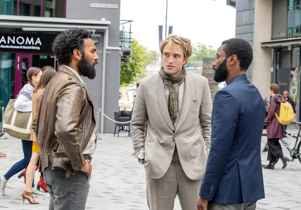 Three men having a conversation on a city street; two wear casual jackets, and one in a light suit with a scarf