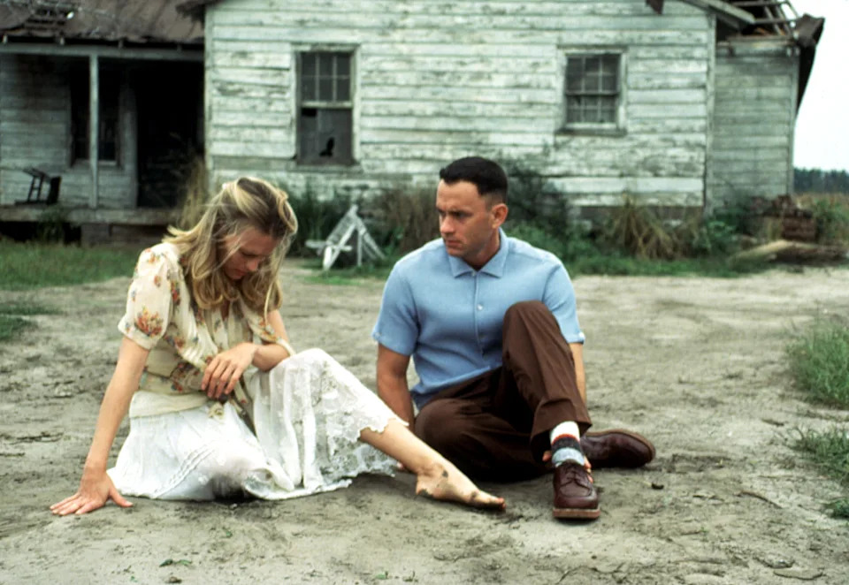 Scene from a film with a man and woman sitting on the ground. The man is in a short-sleeved shirt and slacks; the woman is in a floral blouse and skirt