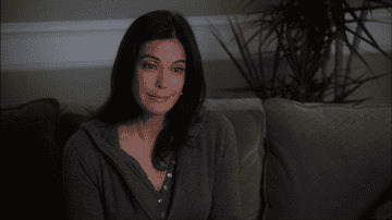 A person sitting on a couch looks towards the camera, wearing a casual, dark, long-sleeve top
