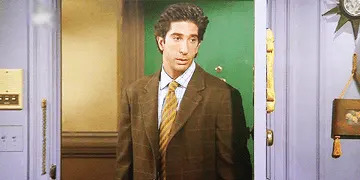 Person wearing a suit and tie stands in a doorway, looking sideways with a neutral expression. Indoors, with visible wall decor