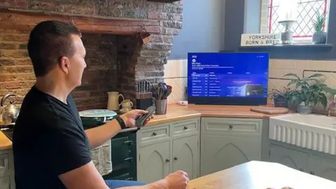 BBC A man sitting in front of a blue screen TV in a kitchen. He is holding a remote in his left hand and is wearing a black T-shirt and blue jeans. 

A sign above the TV reads 'Yorkshire Born & Bred'.

A knife block, several plants and a fruit bowl are visible in the kitchen.
