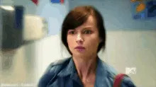 A person with short hair looks shocked or surprised while walking in a hallway, wearing a denim jacket