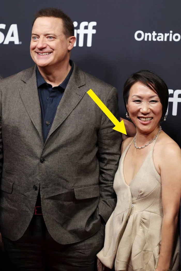 Two individuals smiling on a red carpet. The man wears a tailored suit, and the woman is in a sleeveless, fitted gown