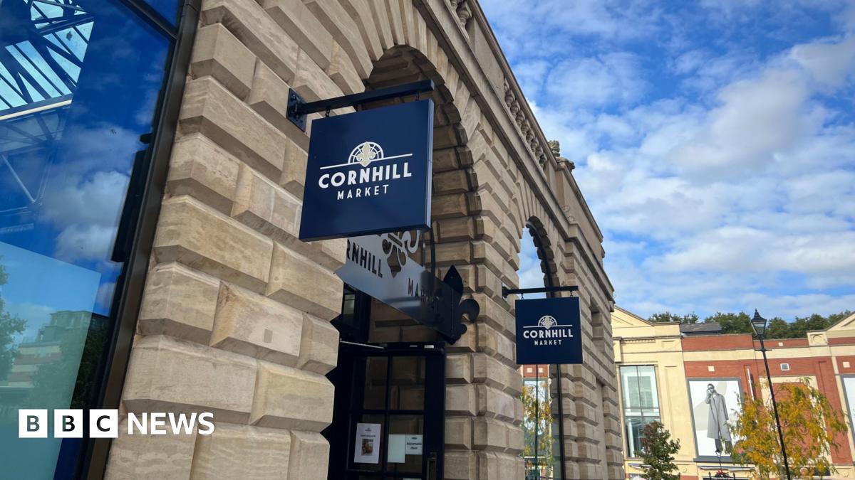 An exterior view of Cornhill Market showing a modern building with navy signs hanging above the arched entrance reading 'Cornhill Market'. The sky is blue with light clouds.