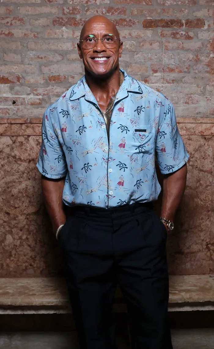 Smiling man in a short-sleeve patterned shirt, black pants, and loafers stands against a textured brick wall
