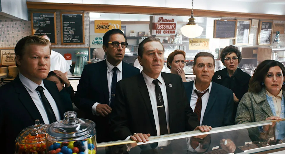 A group of actors in 1950s-style suits and dresses stand behind a diner counter, appearing shocked and intrigued