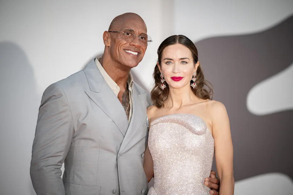Two people are posing on a red carpet. One is wearing a light-colored suit, the other a strapless, shimmering gown with elegant accessories