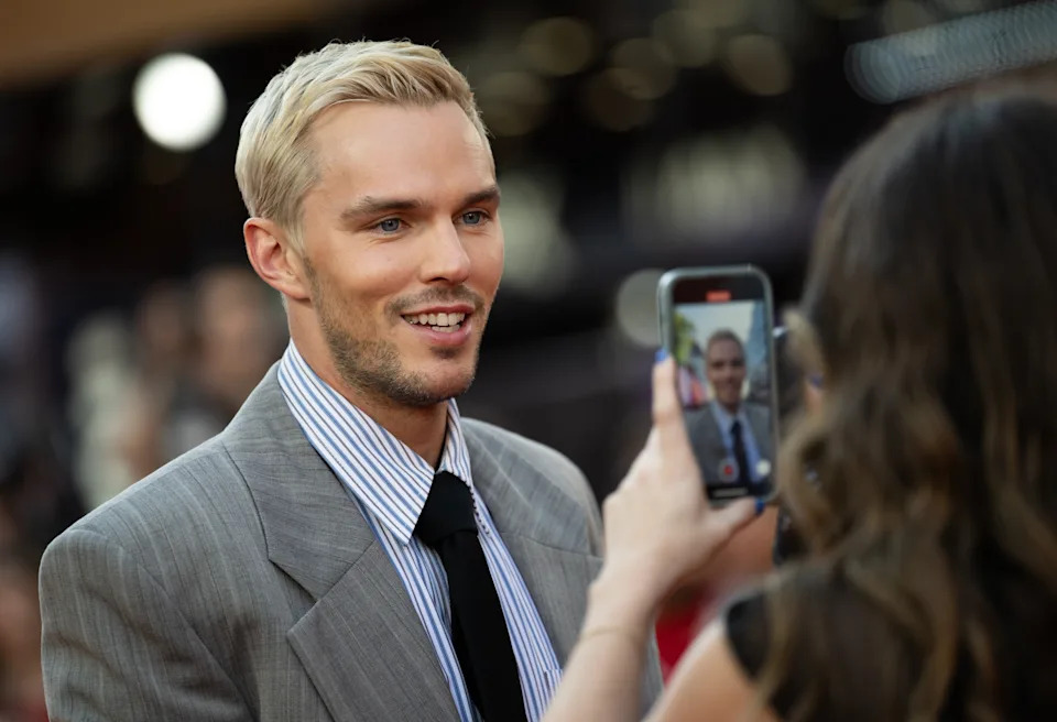 Nicholas Hoult in a suit with wide lapels smiles while someone photographs them with a phone at an event