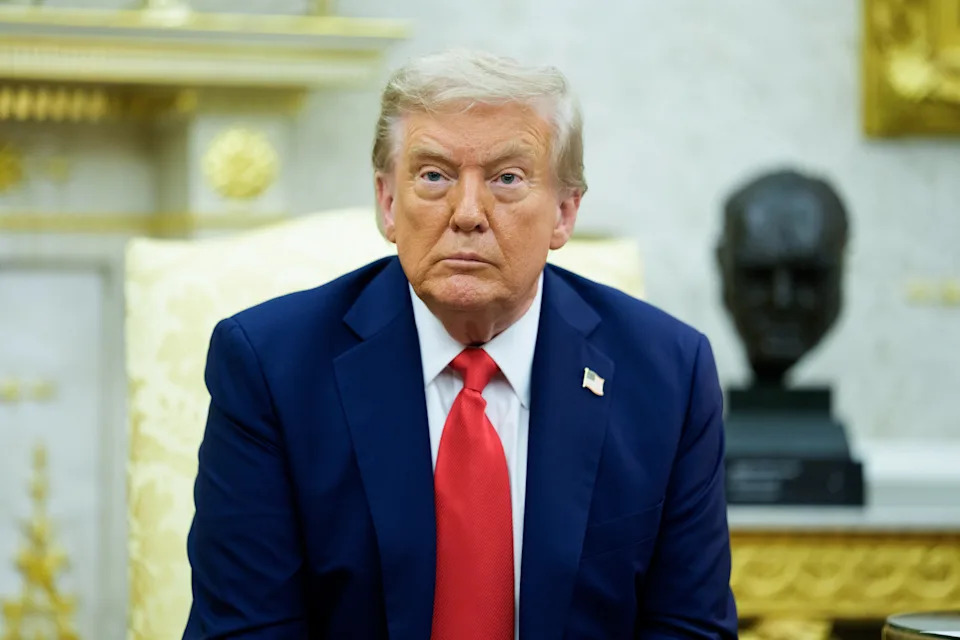 A person in a suit and red tie sits in a formal setting, with a sculpture visible in the background
