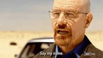 Man in glasses and jacket sternly says "Say my name" at camera in desert setting, with a car in the background